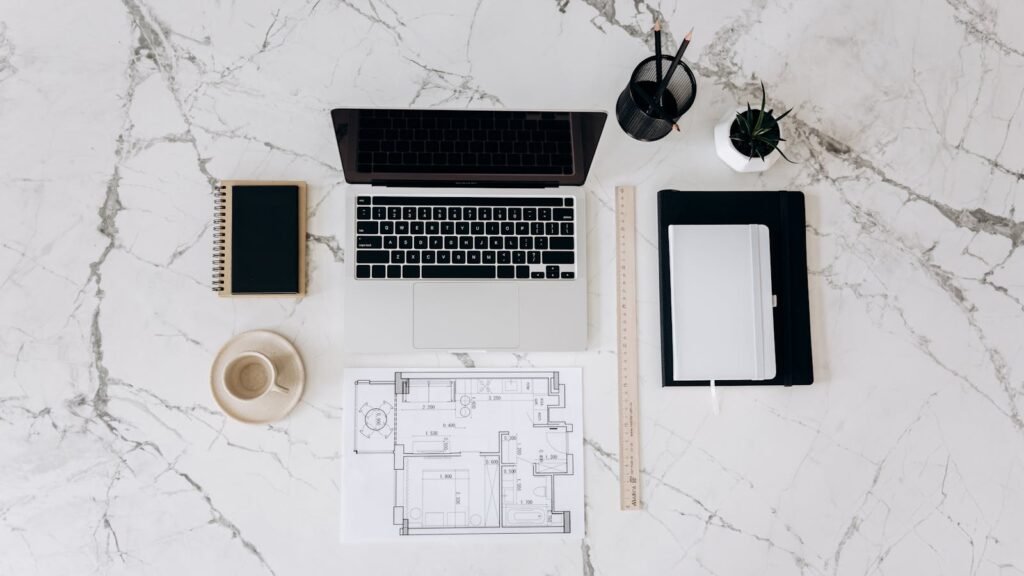 Flat lay of a clean and organized workspace with a laptop, blueprint, and minimal accessories on a marble table.